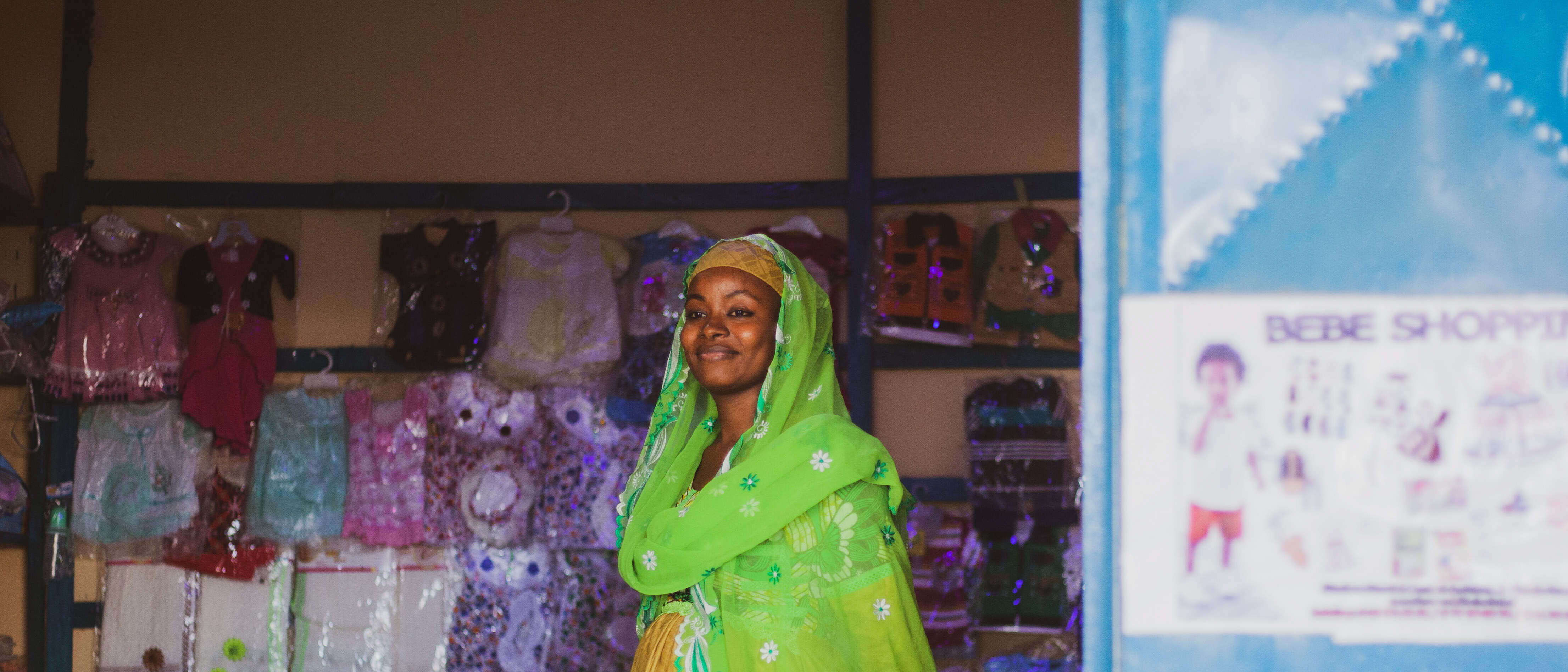 A woman, wearing a vibrant green headscarf and yellow dress, smiles in a shop in Cameroon.
