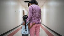 A Mexican mother wearing pink pants and a purple shirt stands with her arms around her young daughter, who has a long braid, in a hallway. They are asylum seekers in an IRC welcome center.